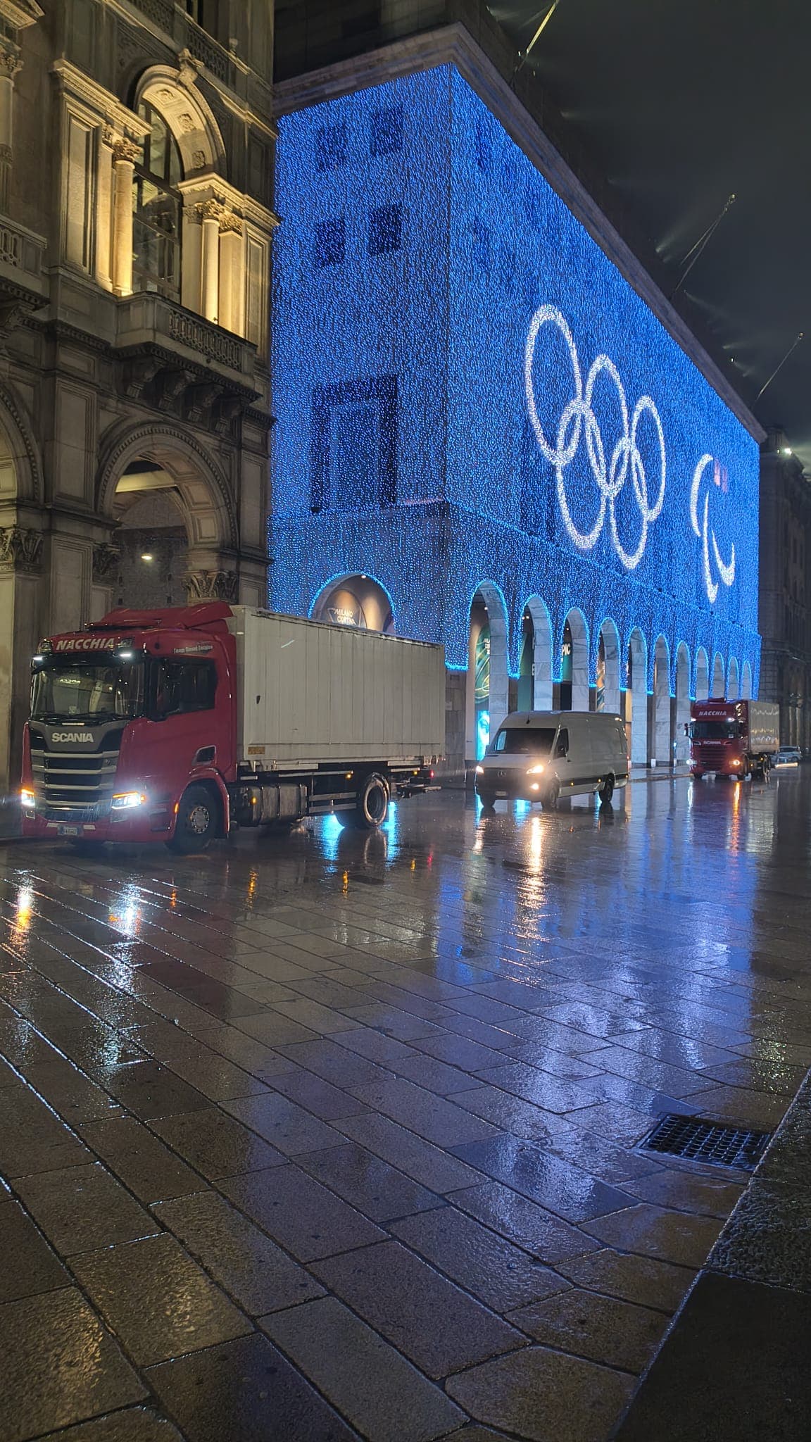 Red truck on wet night street beside a building illuminated with blue Olympic rings.