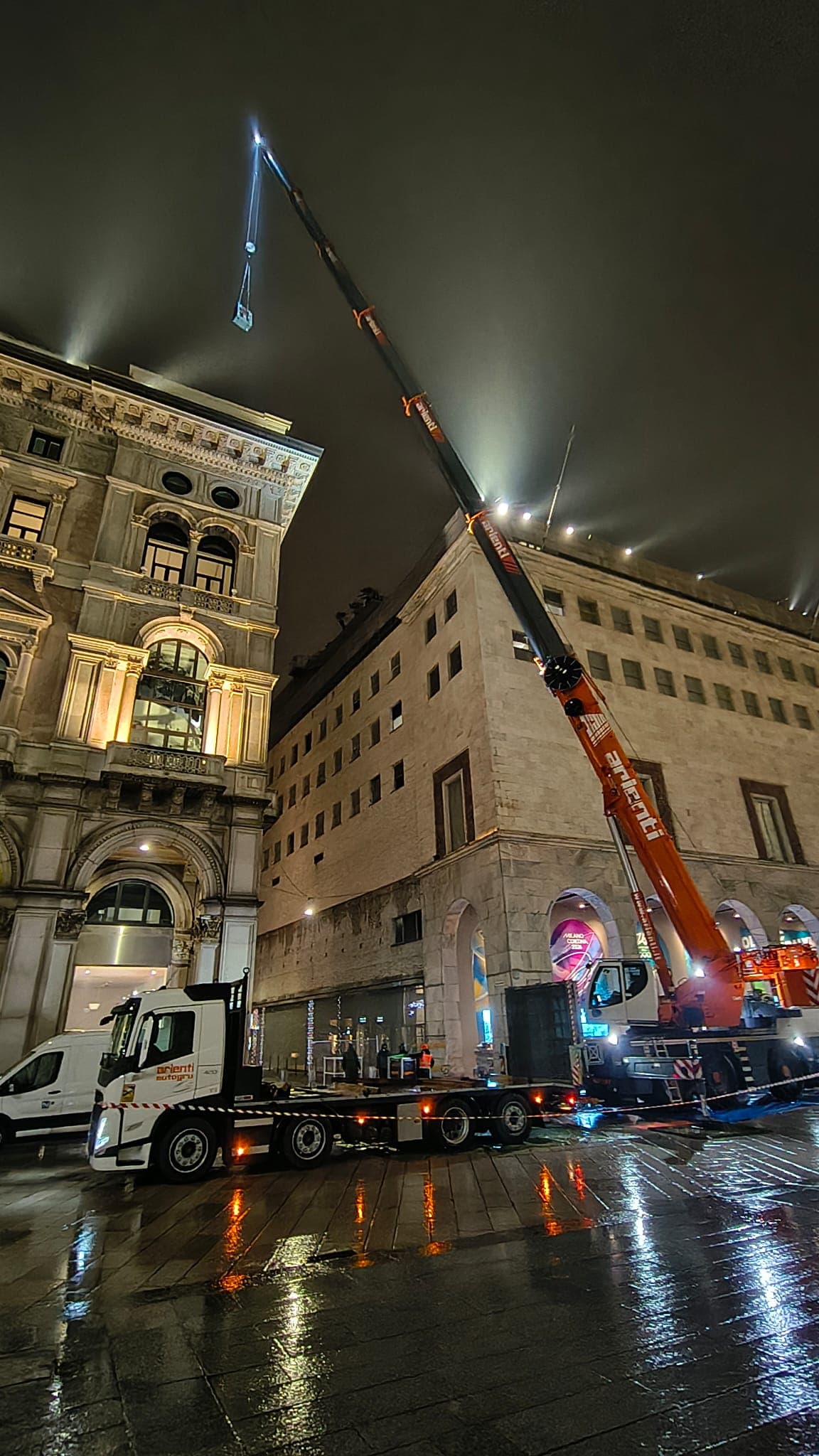 Orange crane lifting a load to a building roof on wet city streets at night.