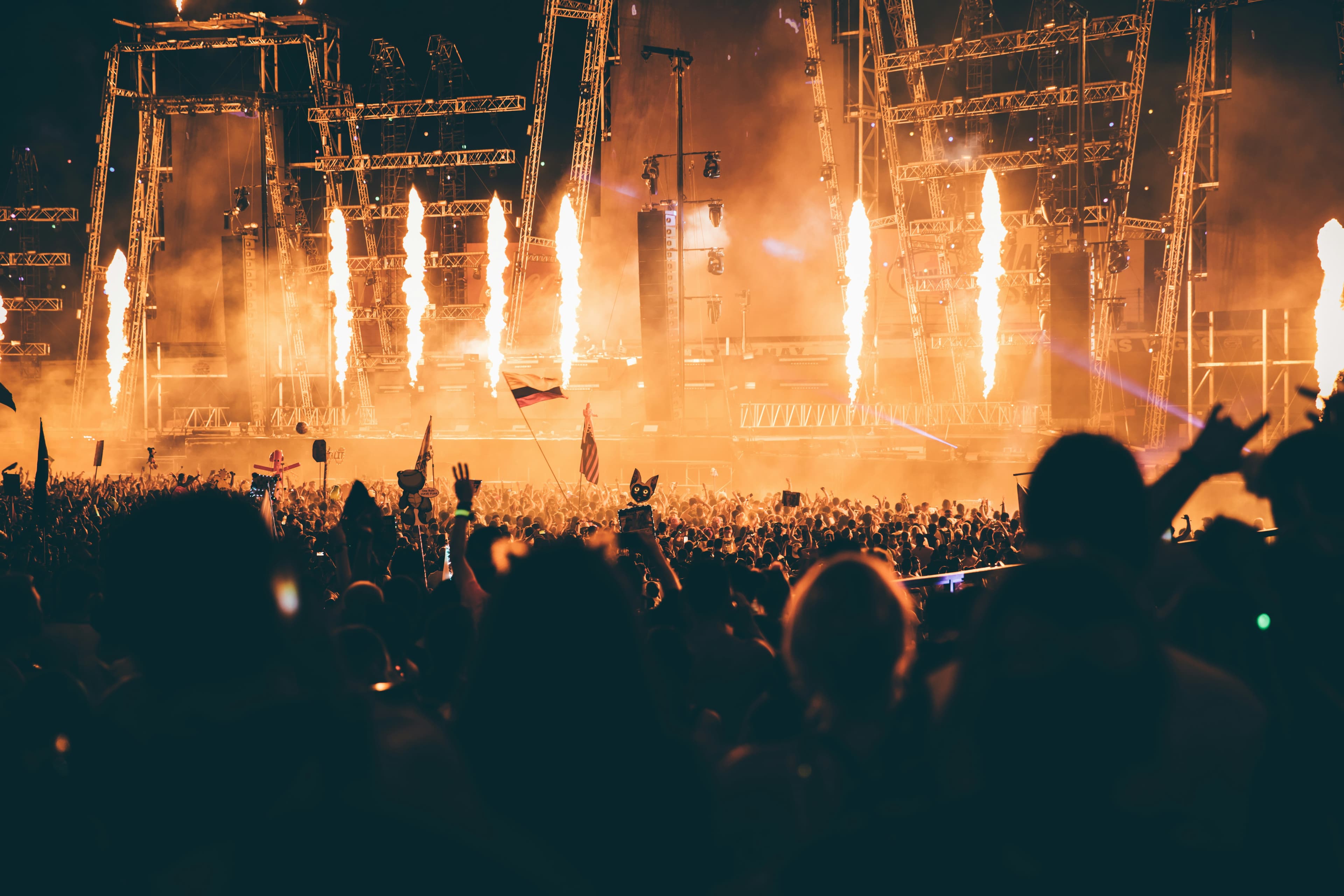 Silhouetted crowd at a music festival with vertical fire jets erupting from a massive stage.