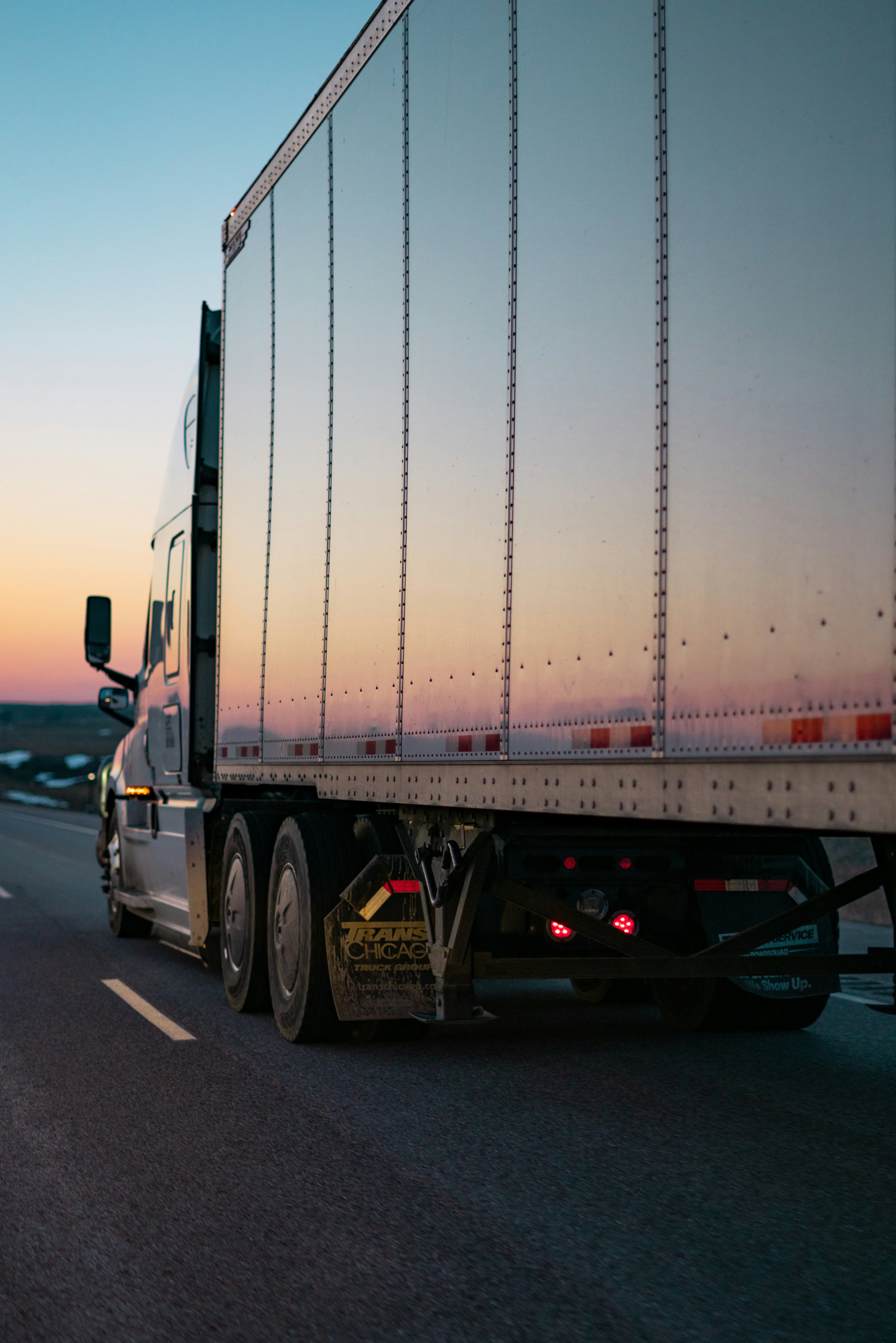 Semi-truck driving on a highway at sunset, its silver trailer reflecting the colorful sky.