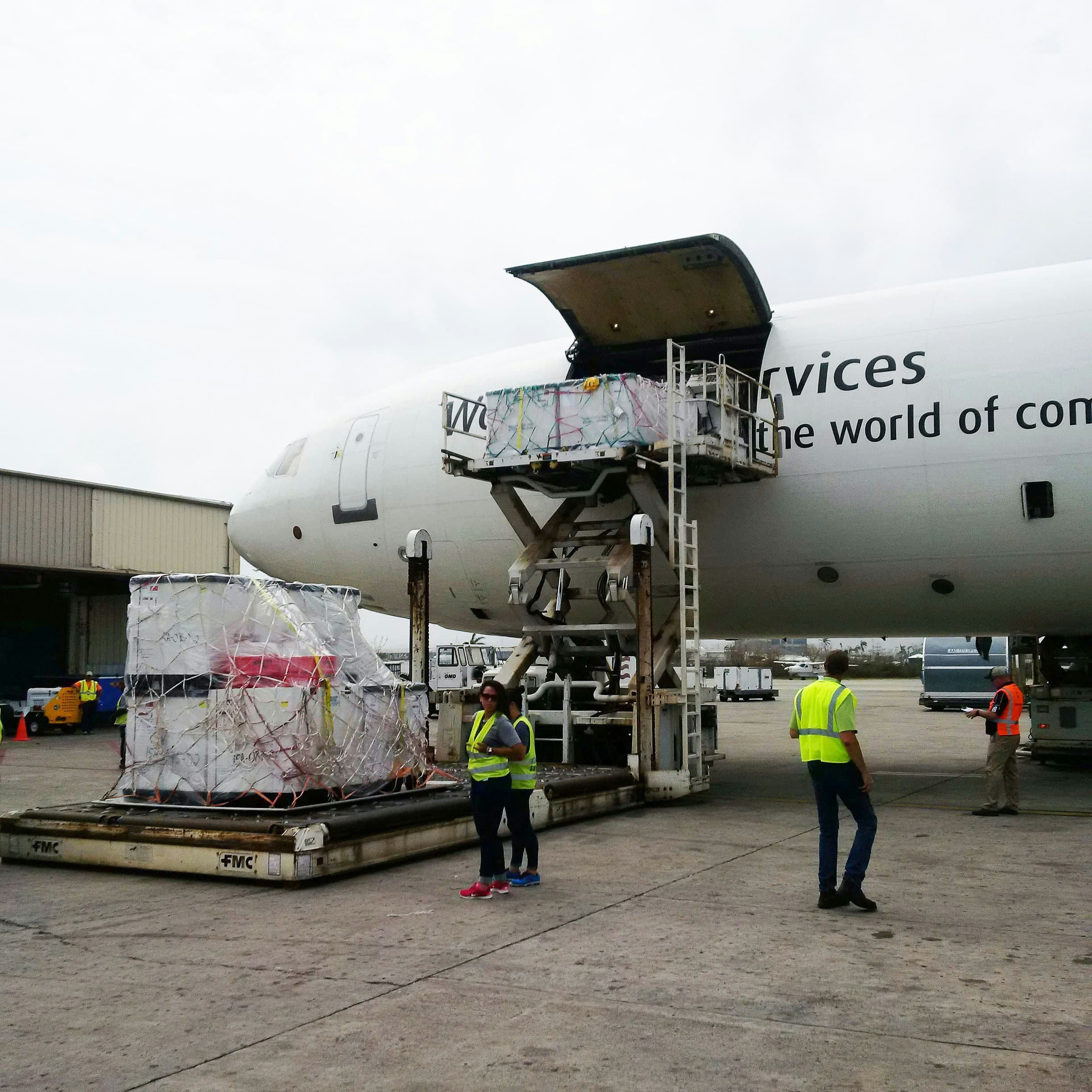 Ground crew loading large wrapped cargo pallets into a white airplane using a mechanical lift.