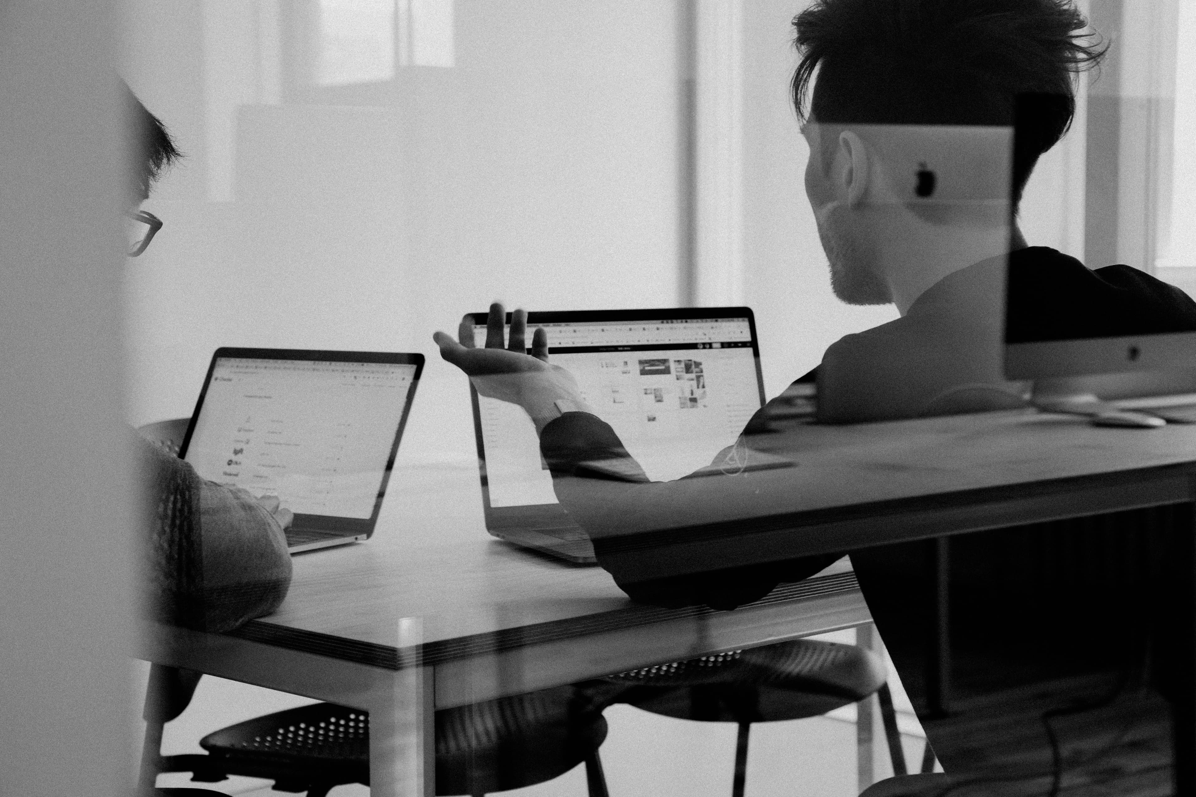 Two people collaborating on laptops in a black and white office, seen through reflective glass.