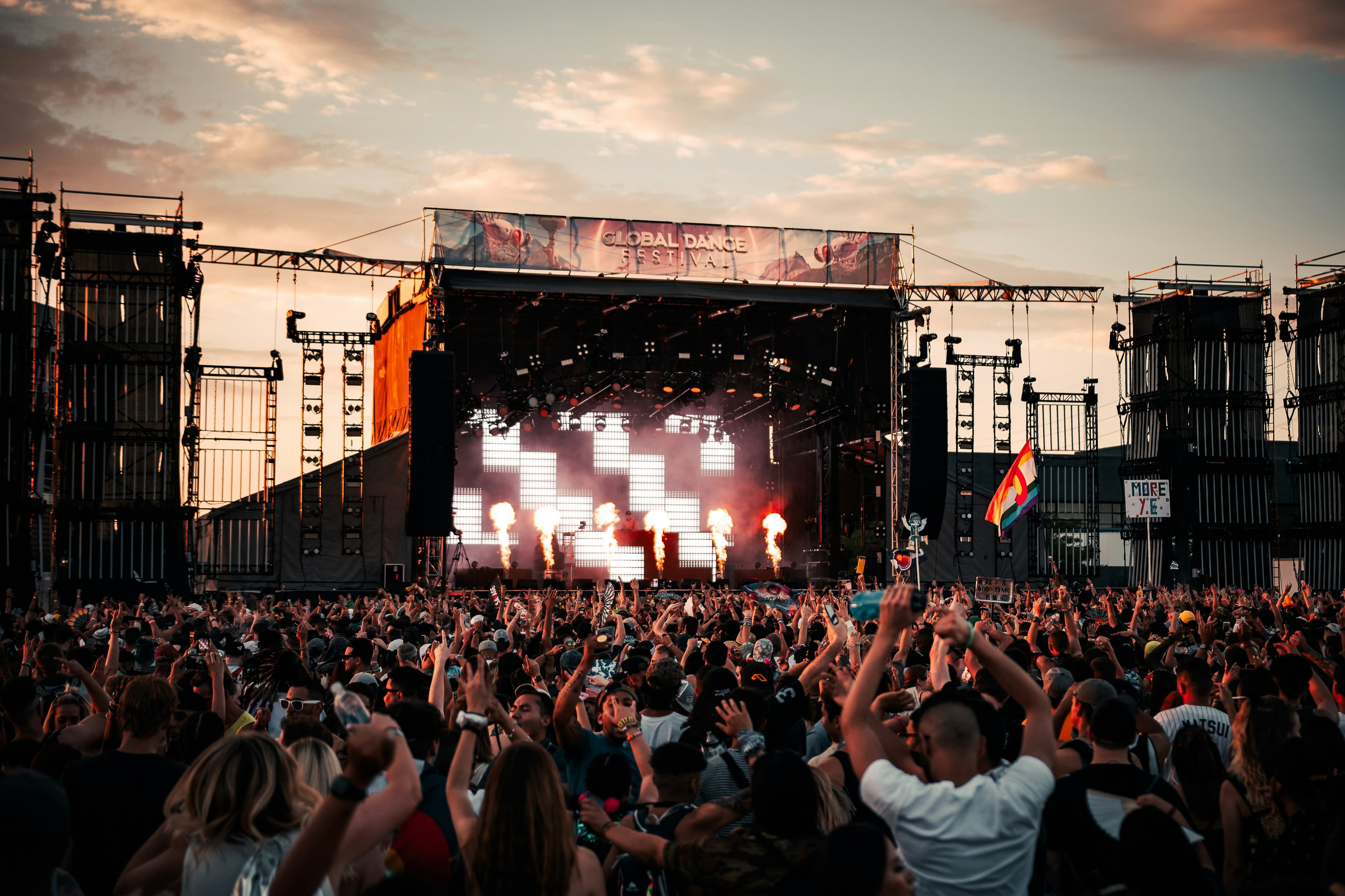 Crowds cheer at Global Dance Festival with fire pyrotechnics on stage under a sunset sky.