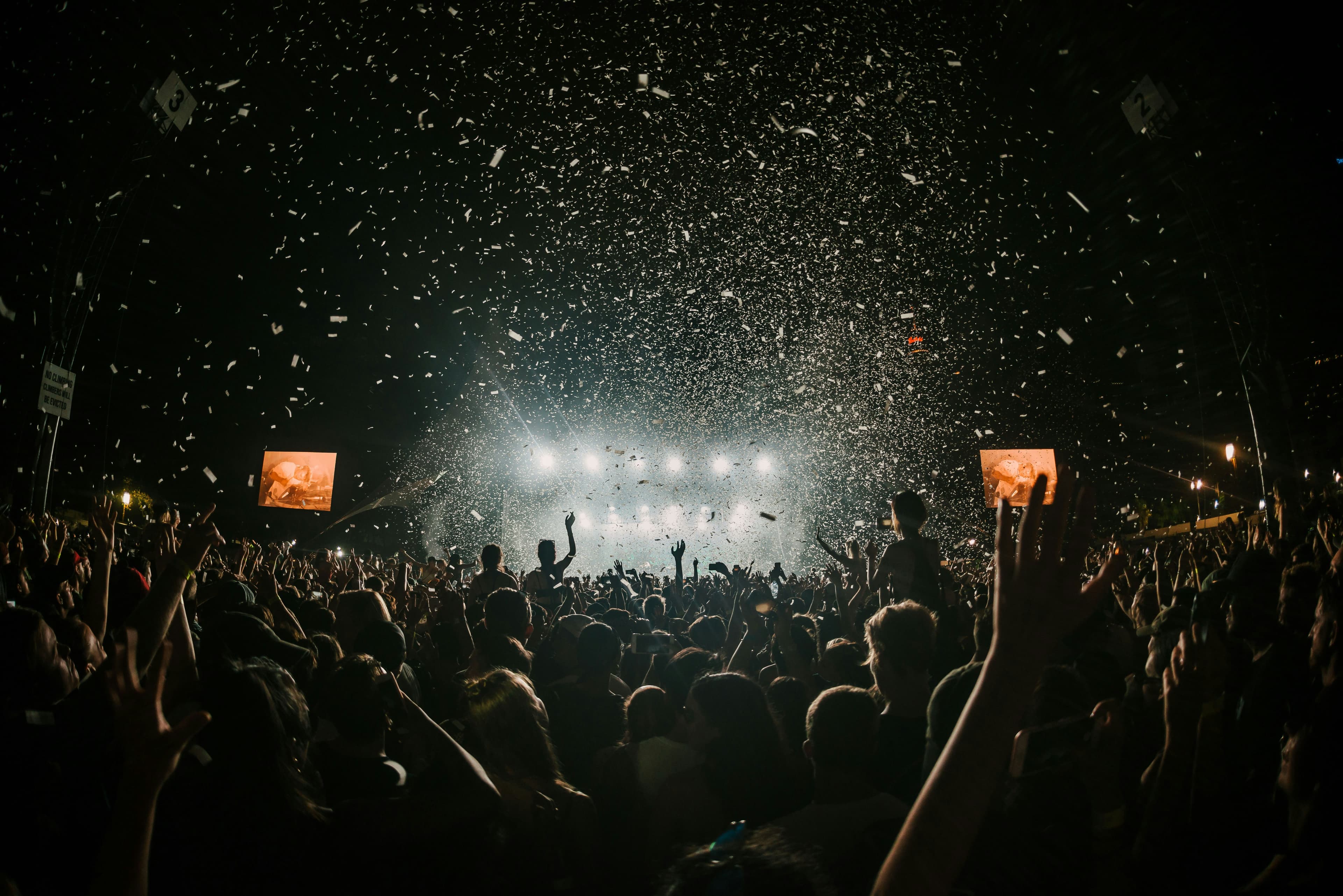 Nighttime concert crowd with raised hands under bright stage lights and falling white confetti.