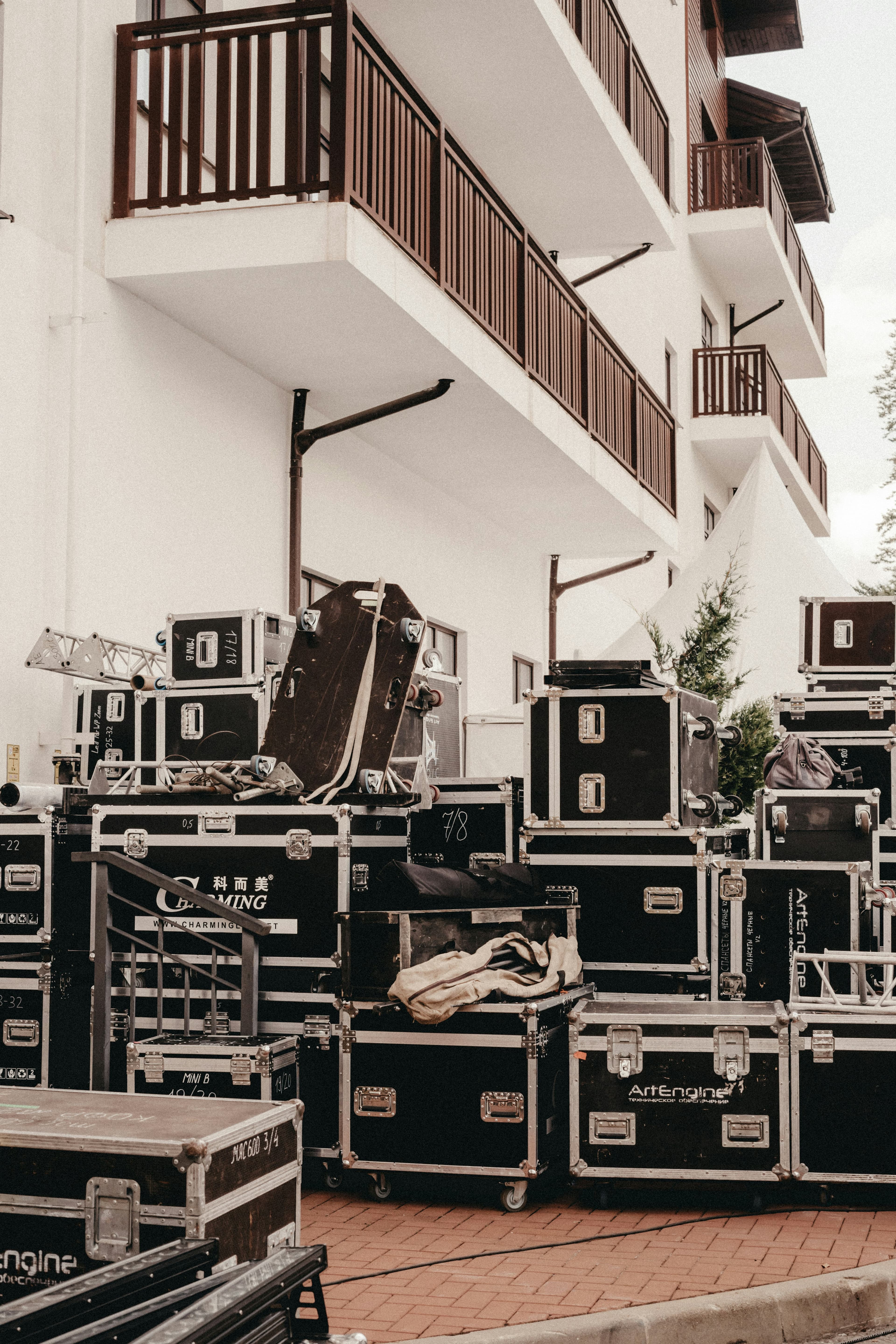 Stacked black road cases and production gear outside a white building with wooden balconies.