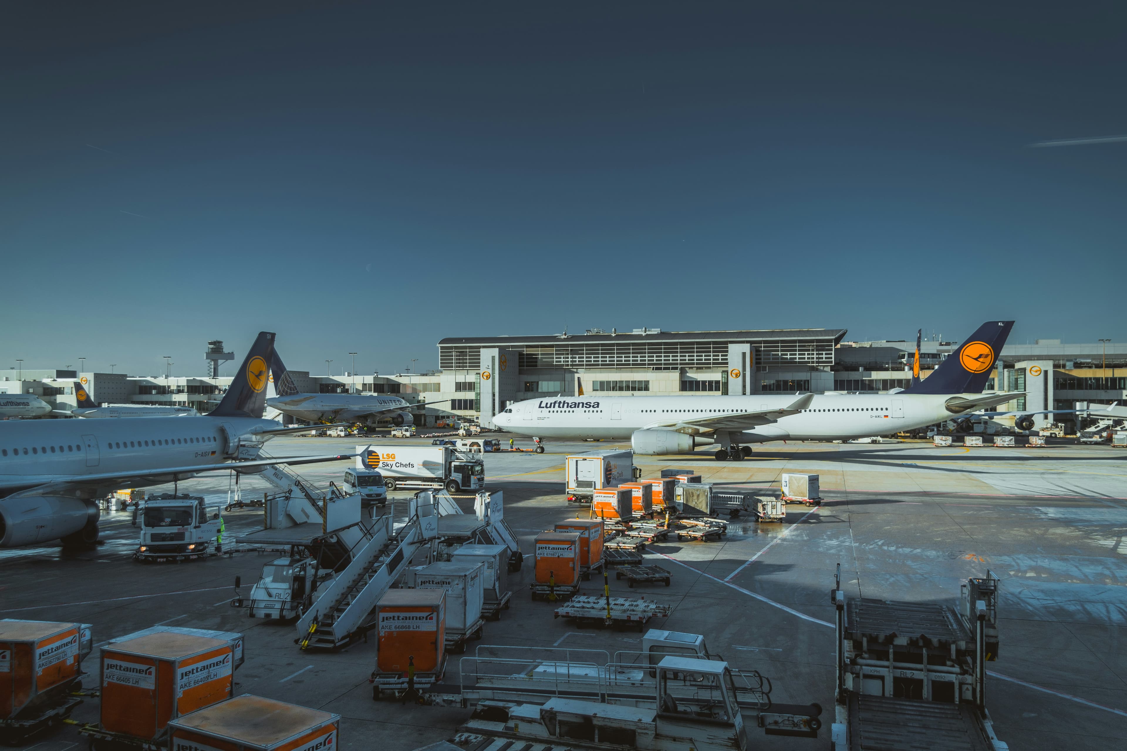 Lufthansa airplanes parked at a busy airport terminal with ground service vehicles and cargo containers.