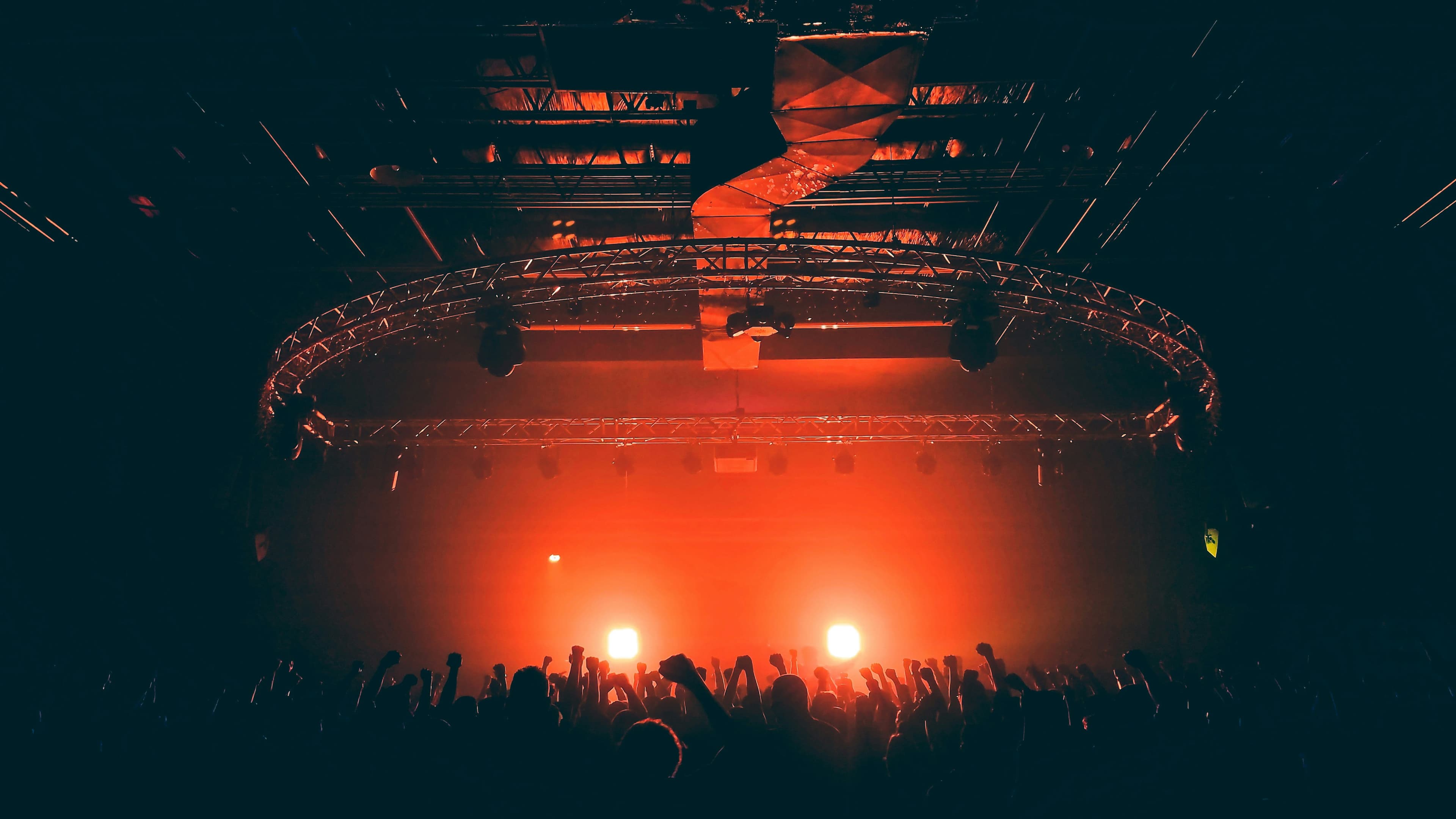 Silhouetted crowd with raised hands under intense orange stage lighting and a circular rig.
