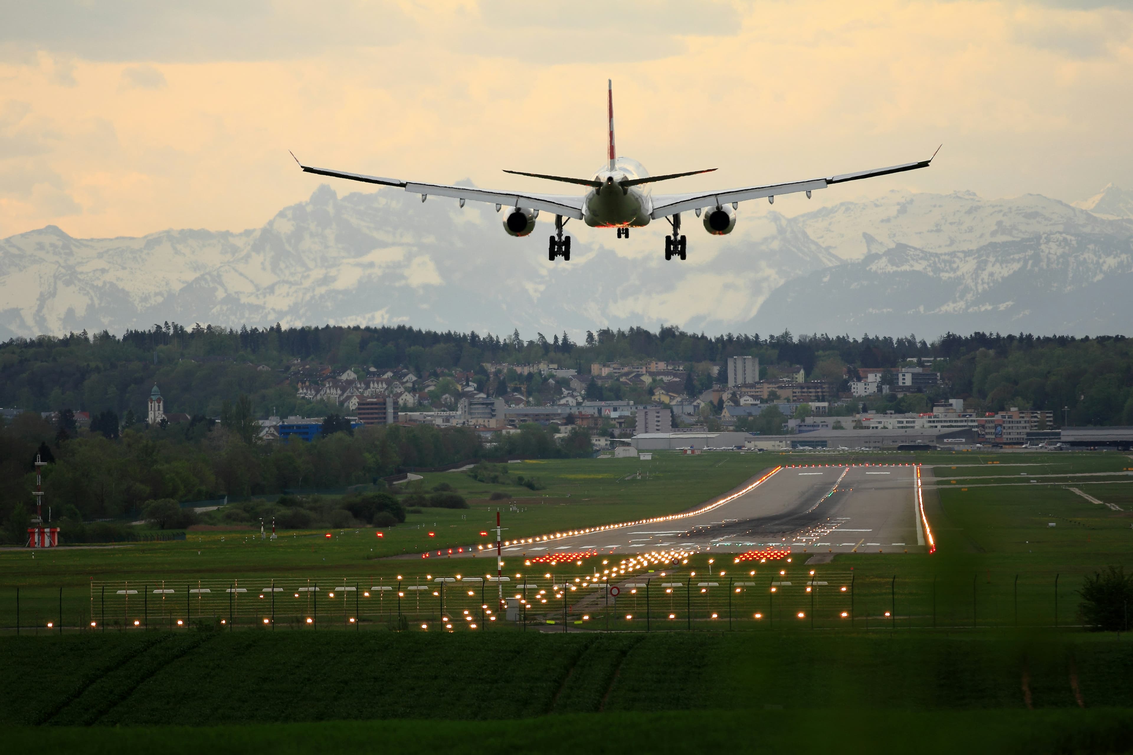 Airplane landing on a lit runway with a town and snowy mountains in the background.
